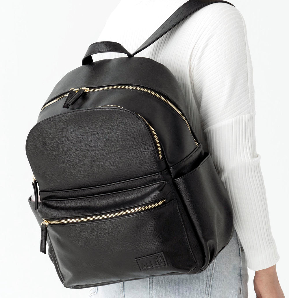 A person wearing a white ribbed top and carrying a Backpack Changing Bag with gold zippers and an "ALLIS" logo embossed at the bottom